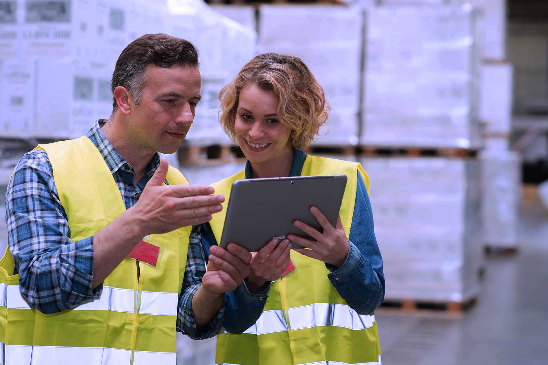 Two warehouse employees in high-visibility vests use a tablet Warehouse management